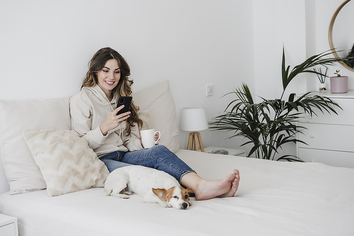 Young woman with coffee mug using phone sitting by dog on bed at home, Photo by Eva Blanco