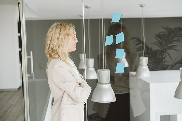 Businesswoman looking at adhesive notes on glass wall in office, Photo by Eva Blanco