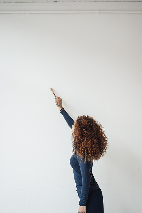 Young woman with curly hair using paintbrush on white wall, Photo by Joseffson