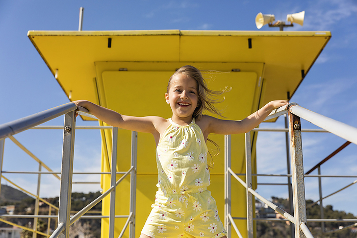 Cheerful girl standing in front of yellow lifeguard hut on sunny day, Photo by Mertxe Alarcón