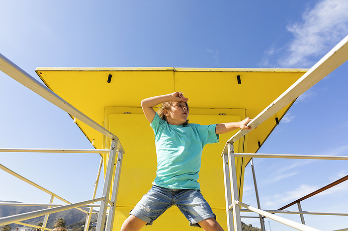 Boy shielding eyes standing amidst railings in front of yellow lifeguard hut, Photo by Mertxe Alarcón