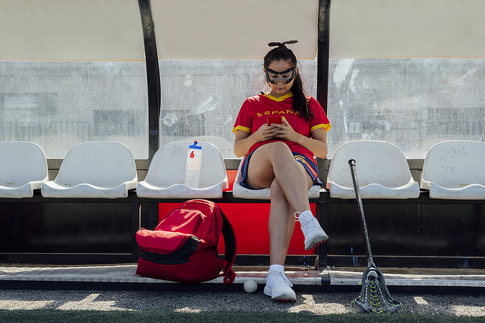 Young lacrosse player using mobile phone on bench at dugout, Photo by Miguel Partido