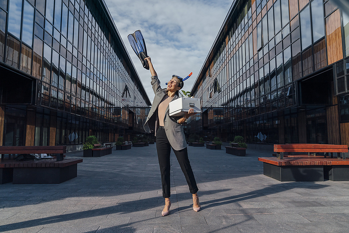 Happy businesswoman with hand raised holding diving flipper in front of office building on sunny day, Photo by Vasily Pindyurin