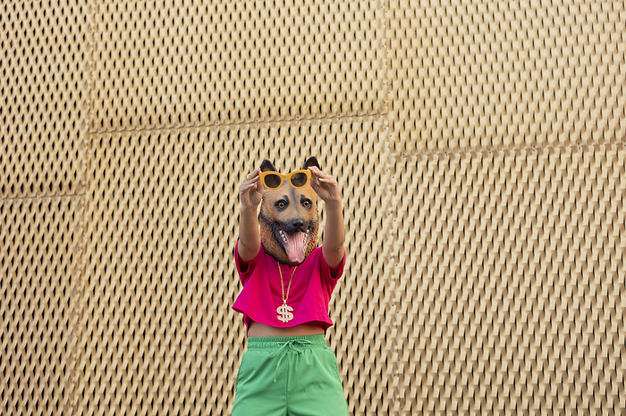 Young woman wearing dog mask holding sunglasses in front of wall