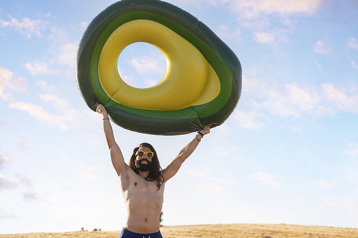 Smiling hipster man with hand raised holding inflatable ring on sunny day