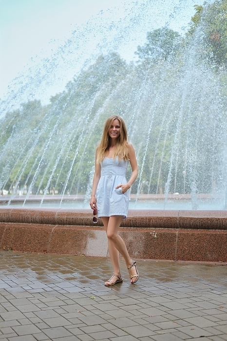 Attractive girl in short blue dress stands in front of fountain in summer park, Photo by Aleksei Isachenko