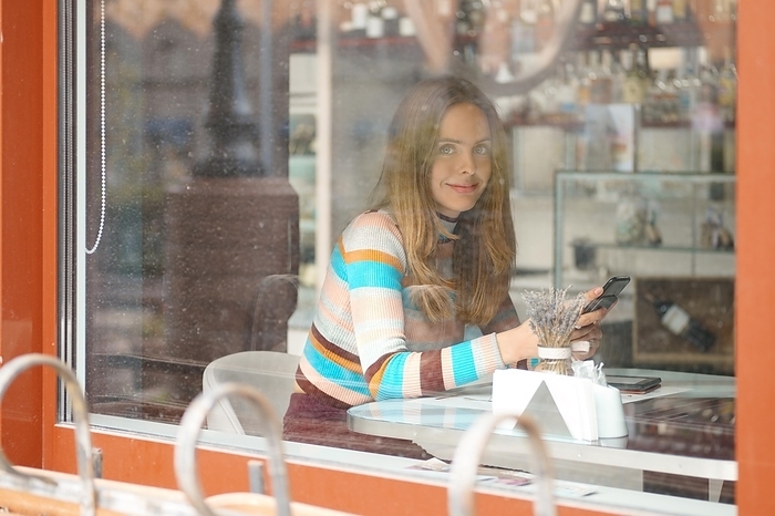 Cute girl with long hair sitting in caffe and chatting on mobile phone. Photo taken from outside, with a see through the window, Photo by Aleksei Isachenko