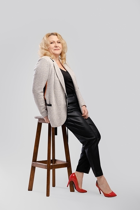 Portrait of senior woman in leather trousers and blazer sitting in profile on over grey studio background, Photo by Aleksei Isachenko
