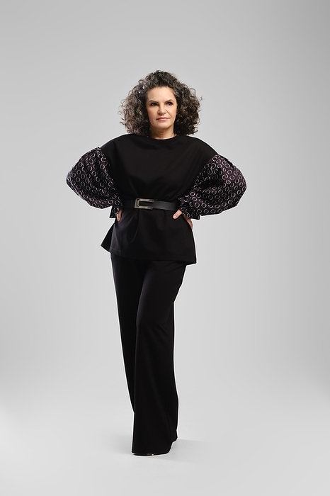 Senior woman with lush curly hair and black pantsuit posing in studio with hands on waist over grey background, Photo by Aleksei Isachenko