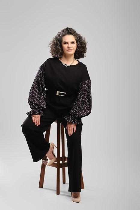 Senior woman with lush curly hair and black pantsuit sitting on tall chair in studioo over grey background, Photo by Aleksei Isachenko