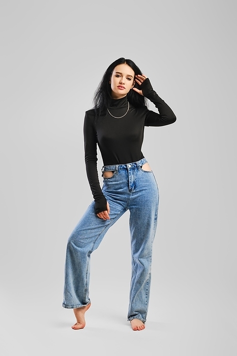 Young barefoot woman posing in studio in black bodysuit with long sleeves and boyfriend jean over grey background, Photo by Aleksei Isachenko