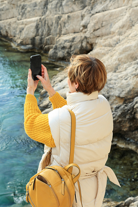 Elderly tourist woman taking photo on rocky sea coast in Greece