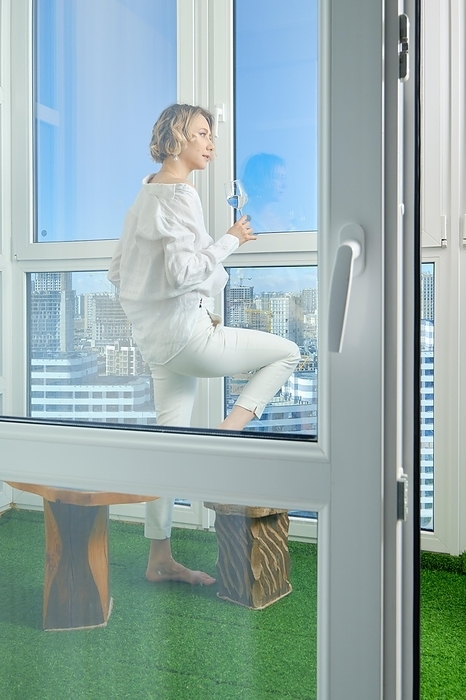 Barefoot woman stands on the balcony of her new apartment and admires the view of the city from the window, Photo by Aleksei Isachenko