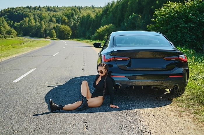 Sexy racer woman in bodysuit and rough boots sits on asphalt and leaning to her powerful racing car, Photo by Aleksei Isachenko