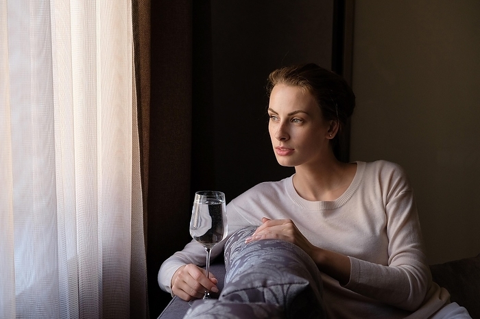 Attractive girl with glass of water in hand sitting at home on the sofa and looking to the window, Photo by Aleksei Isachenko