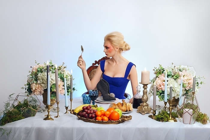 Fashion blonde model with colorful makeup sitting behind festive decorated table and eating felt shoe, place for text, Photo by Aleksei Isachenko