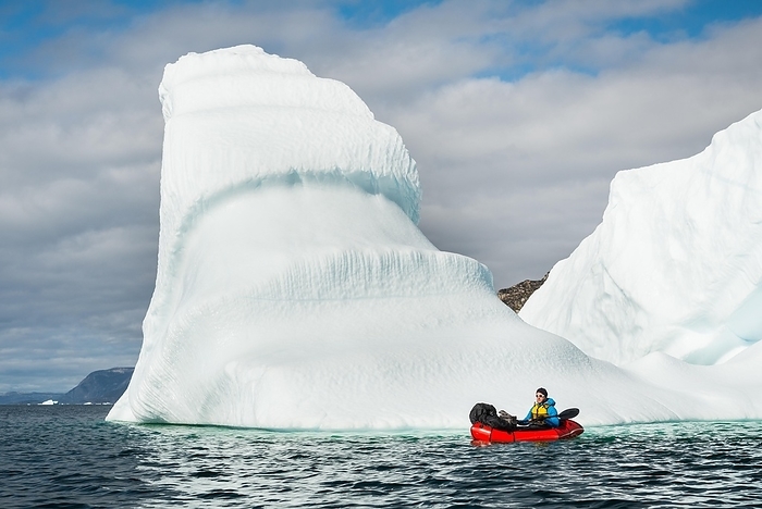 Man with packraft on fjord, icebergs and mountains behind, partly cloudy, Greenland, North America, Photo by Gabriel Gersch