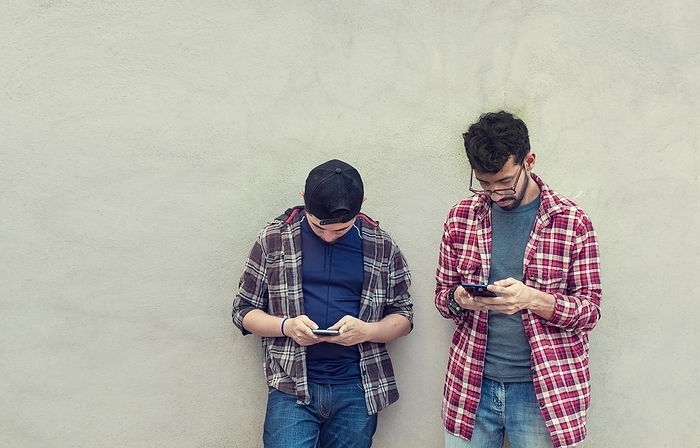 Two teenage friends on a wall checking their cell phones, Two friends leaning on a wall texting on their phones. Friend showing cell phone to his friend, Smiling friends checking cell phones, Photo by Isai Hernandez