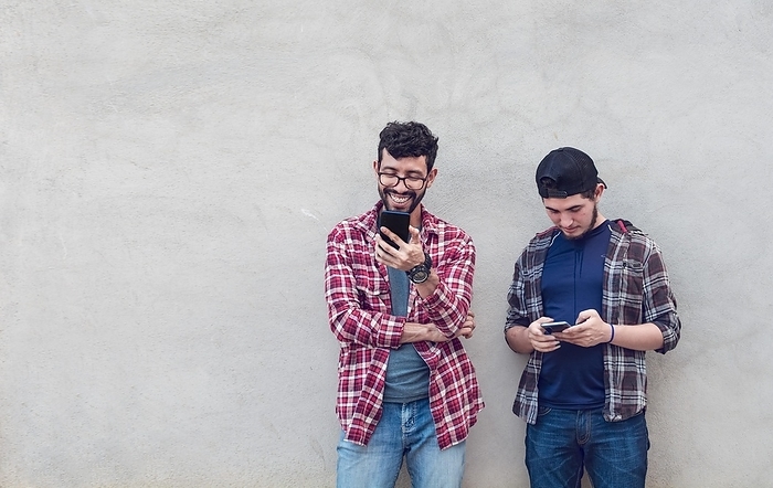 Two smiling friends leaning on a wall checking their cell phones, Friends leaning on a wall texting on their phones. Friend showing cell phone to his friend, Smiling friends checking cell phones, Photo by Isai Hernandez