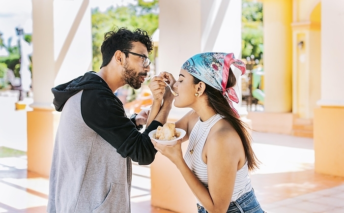 A couple eating shaved ice in a square, Young couple eating shaved ice together with arms intertwined, Side view of young couple with arms linked eating ice cream in a city square, Photo by Isai Hernandez