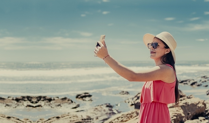 Young woman on vacation taking photos on the beach, Smiling girl in hat taking photos on the beach. Beach vacation concept. Girl in hat on the beach taking a selfie, Photo by Isai Hernandez