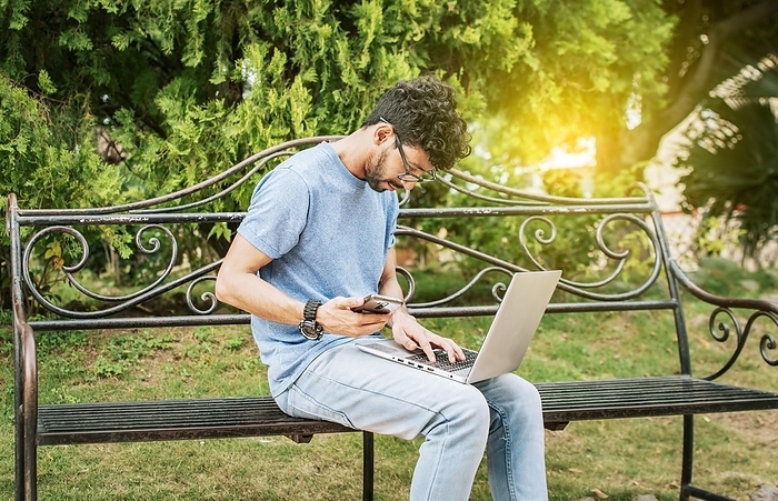Young man in a park with laptop and cell phone. Freelancer man sitting in a park using laptop and cellphone. Man in a park working online with laptop. Relaxed man working with laptop outdoor, Photo by Isai Hernandez