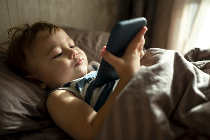 Cute baby boy using smart phone in duvet in bed at home
