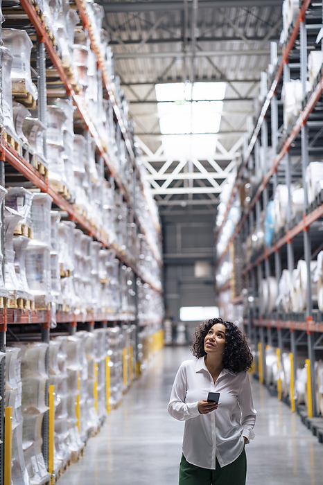 Businesswoman holding smart phone standing with hand in pocket at factory