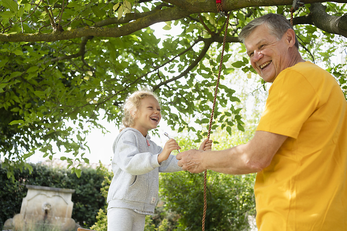 Happy senior man with granddaughter making swing with rope in garden