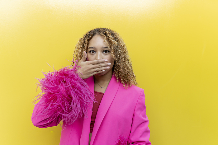 Young woman covering mouth with hand in pink fur blazer against yellow background