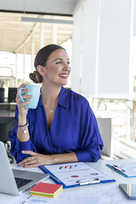 Smiling young businesswoman with coffee cup at desk in office