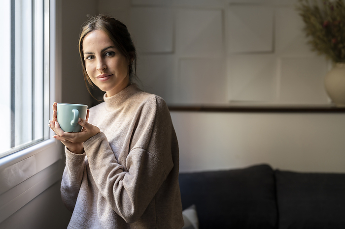 Smiling young woman with cup of tea standing by window at home