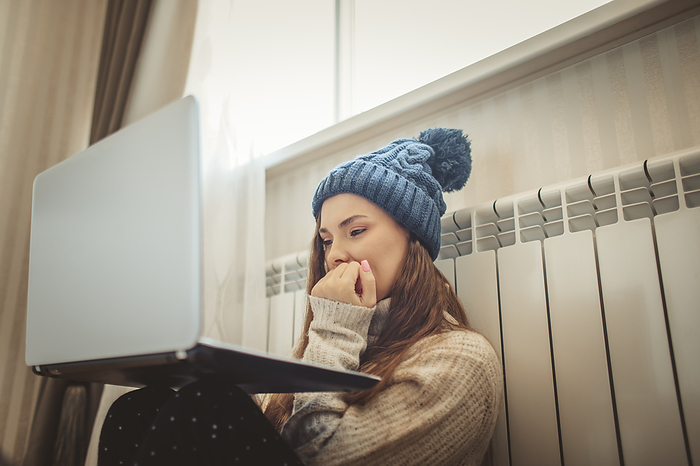 Worried woman wearing knit hat looking at laptop leaning on radiator