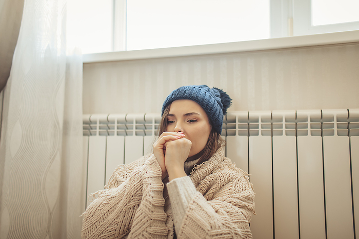 Woman wearing knit hat wrapped in blanket leaning on radiator