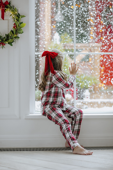 Girl in red pajamas looking snowfall through window at home