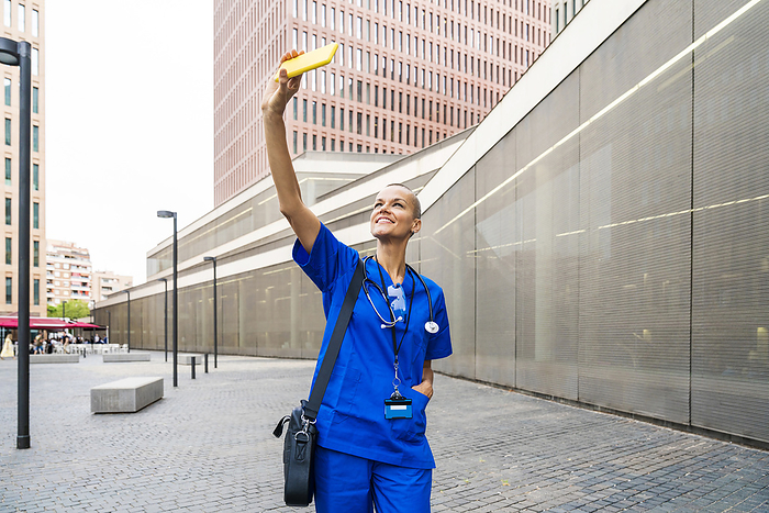 Smiling mature nurse with shoulder bag taking selfie on smart phone