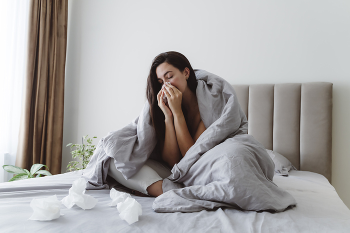 Ill woman blowing nose in tissue on bed at home
