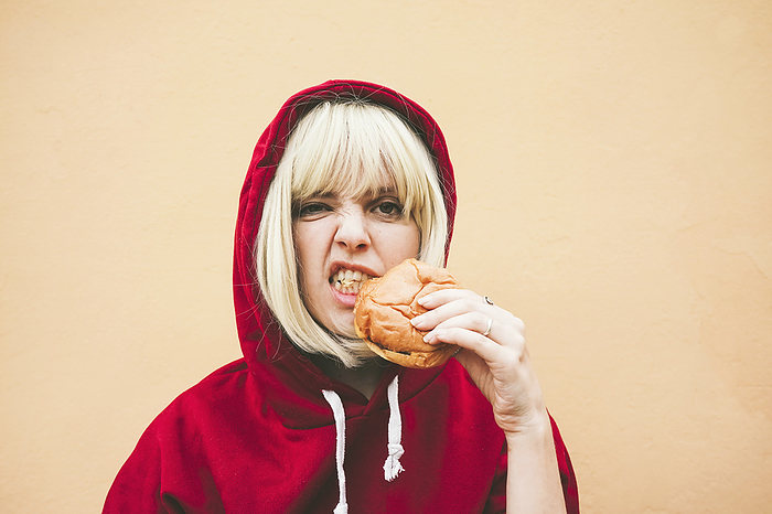 Woman wearing red hooded shirt eating hamburger in front of peach wall