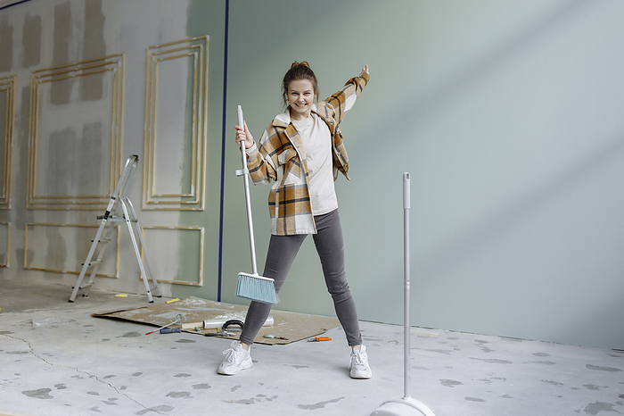 Cheerful woman in plaid shirt dancing with broom in apartment