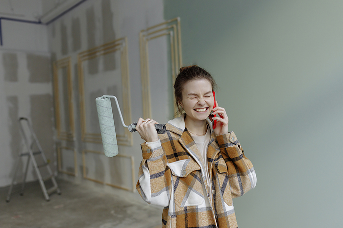 Happy young woman holding paint roller and talking on smart phone with eyes closed at home
