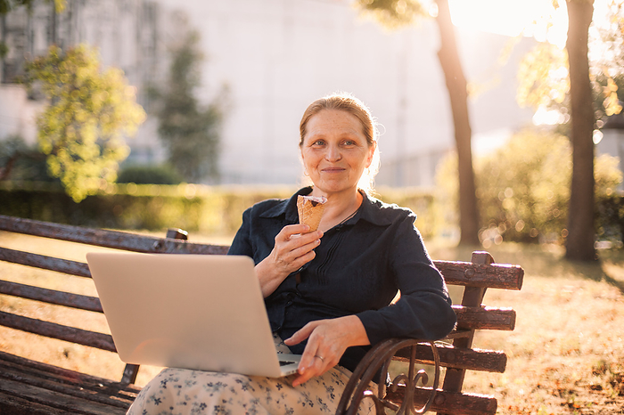 Happy senior business woman holding laptop and ice cream in park