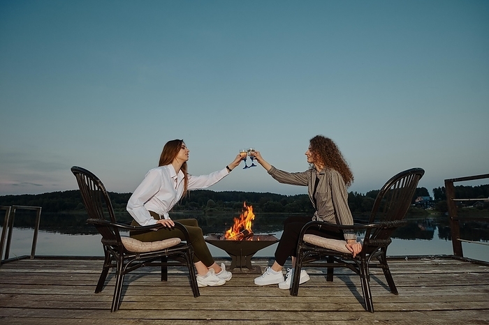 Happy women toasting white wine sitting on pier near the fire