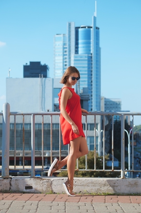 Pretty girl in short silk dress posing under the sun with urban view on background