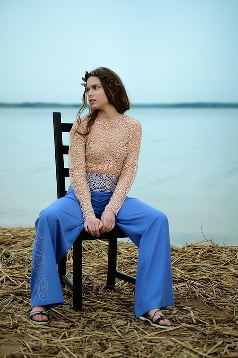 Calm fashion model in wide blue pants and lace blouse sitting on chair on a shore of the lake