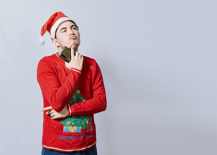 Thoughtful man with christmas hat holding cell phone, Young man in christmas clothes thoughtful with cell phone isolated, Handsome man in christmas clothes thoughtful holding cell phone