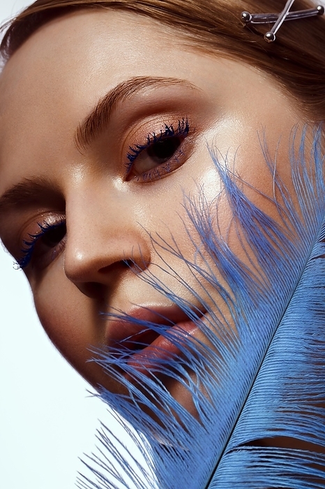 Beautiful tanned girl with creative make-up and blue eyelashes, feather in hand. Beauty face. Photo taken in the studio