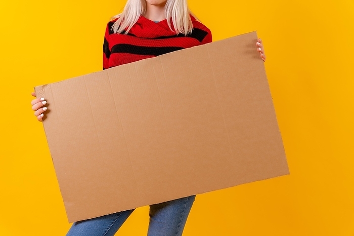 Holding empty cardboard advertisement poster, blonde caucasian girl on yellow background studio