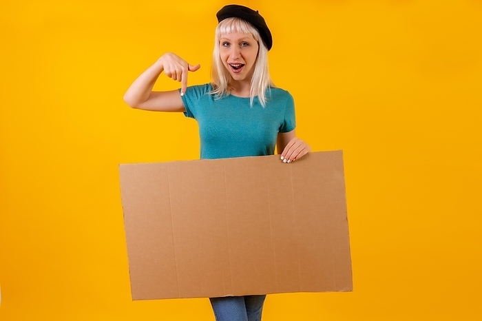 Pointing smiling cardboard advertisement poster, blonde caucasian girl on yellow studio background