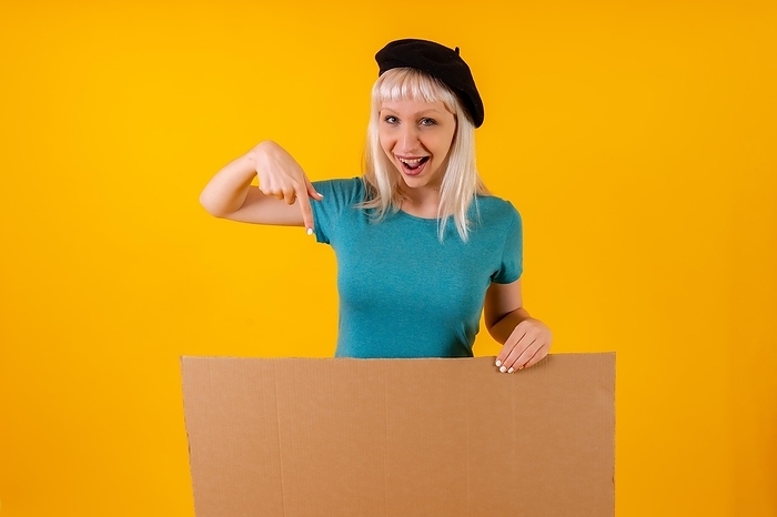 Pointing smiling cardboard advertisement poster, blonde caucasian girl on yellow studio background