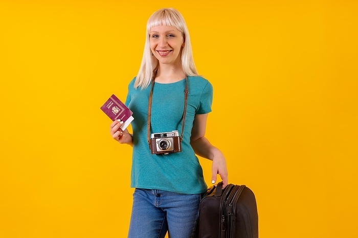 Smiling tourist with suitcase and passport, blonde caucasian girl on yellow background studio